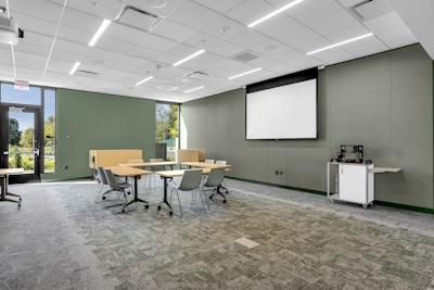 Large room with sage walls and carpet, contains table, chairs, projector screen to accommodate groups