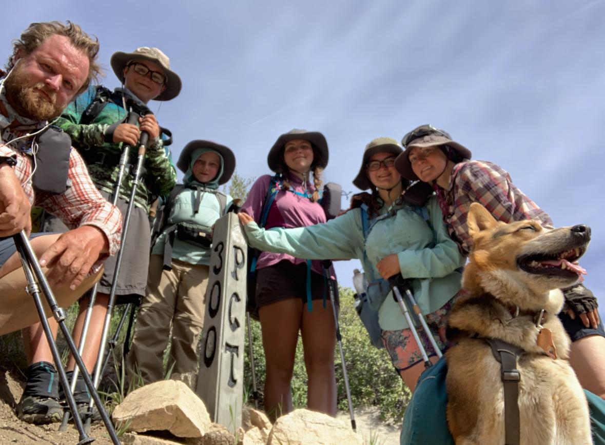 Two adults and four kids dressed in hiking gear pose with their dog on the Pacific Crest Trail under a blue sky.