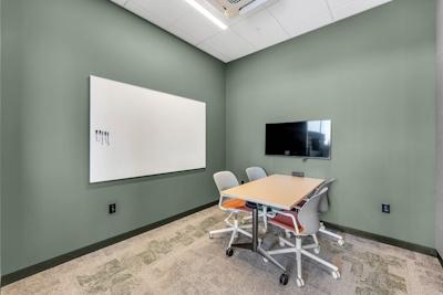 Smaller meeting room with sage green walls, table, chairs, whiteboard, and viewing screen